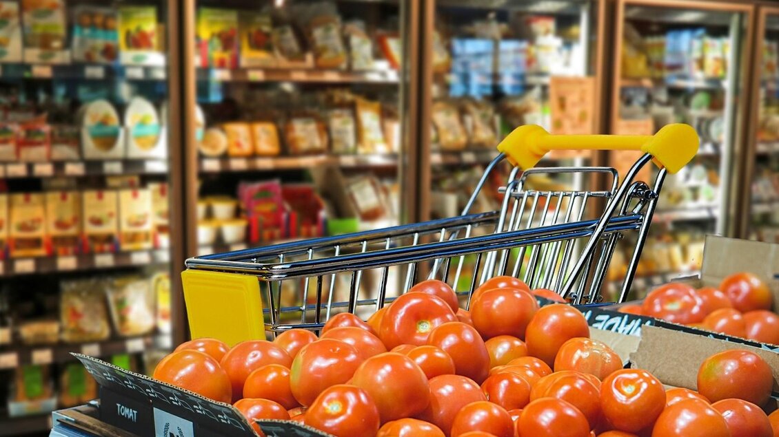Ein Einkaufswagen mit frischen Tomaten steht im Vordergrund. Im Hintergrund sind Regale mit verschiedenen Lebensmitteln zu sehen. | © Symbolfoto Pixabay