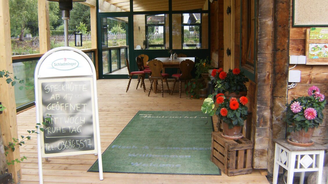 Ein einladender Eingangsbereich eines Cafés mit Holzdekor. Blumen in Töpfen verschönern die Atmosphäre und es sind Tische im Inneren sichtbar. | © Speckhütte
