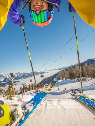 A skier with a colorful helmet and goggles looks down as he descends the slope. In the background, there are snow-covered mountains and clear skies. | © Reiteralm