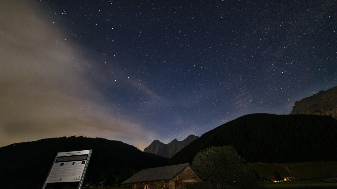 Eine klarer Nachthimmel mit vielen Sternen über einem kleinen, rustikalen Gebäude. Im Hintergrund sind hohe Berge und teilweise bewölkter Himmel zu sehen. | © Gerald Grünwald