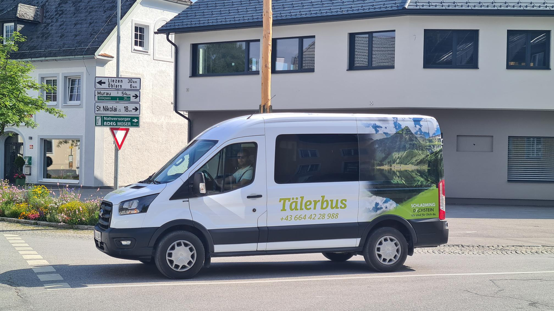 Ein Van mit der Aufschrift „Tälerbus“ steht an einer Straßenecke. Im Hintergrund sind typische Gebäude und Straßenbeschilderungen zu sehen. | © TVB Schladming-Dachstein, Gerhard Piilz