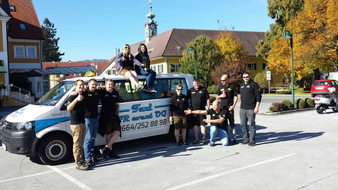 Eine Gruppe von Personen steht um einen Transporter mit dem Schriftzug „Taxi“. Die Szene findet an einem klaren, sonnigen Tag in einer Stadt statt. | © Taxi PR event OG