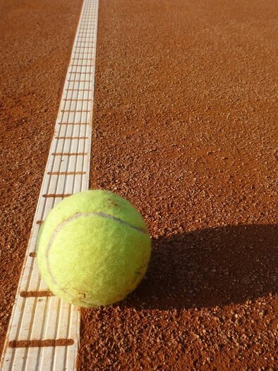 Ein Tennisball liegt auf einem Sandplatz neben der Linie. Der Boden ist rot und die Sonne scheint. | © Hans Dietmann (Pixabay)