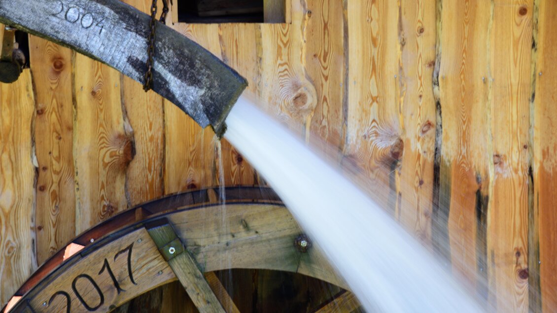 Ein Wasserrad aus Holz mit einem Wasserlauf, der kräftig aus einem Rohr fließt. Im Hintergrund ist eine helle Holzoberfläche zu sehen. | © Naturpark Sölktäler