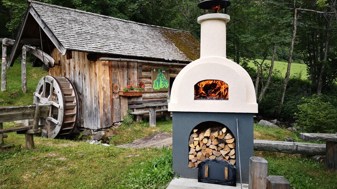 Ein charmantes Holzhaus steht in der Natur, neben einem modernen Kamin mit sichtbarem Feuer. Der Kamin ist mit Holzscheiten gefüllt und bietet eine gemütliche Atmosphäre im Freien. | © Naturpark Sölktäler