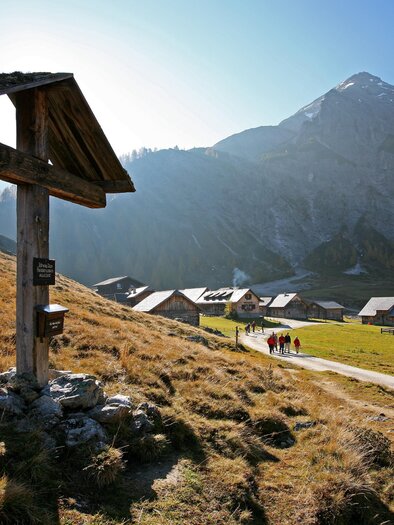 A picturesque mountain landscape with a wooden sign in the foreground. In the background, huts and hikers can be seen on a narrow path. | © Herbert Raffalt
