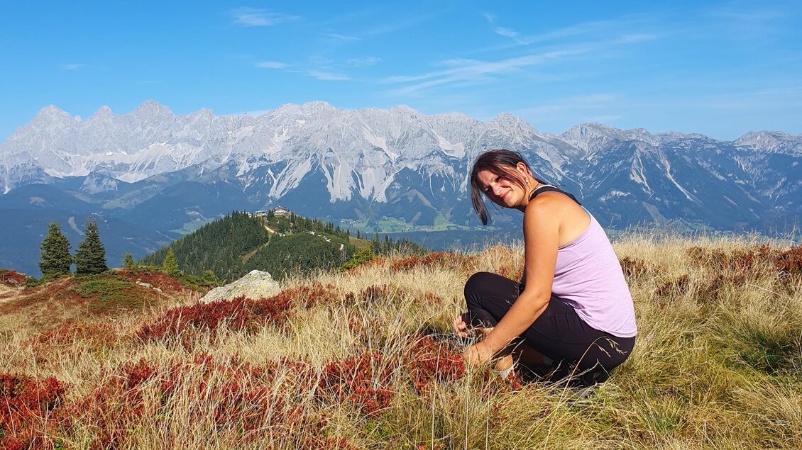 Eine Frau sitzt in buntem Gras auf einem Berg. Im Hintergrund sind majestätische Berge und ein klarer blauer Himmel zu sehen. | © Verena Novak