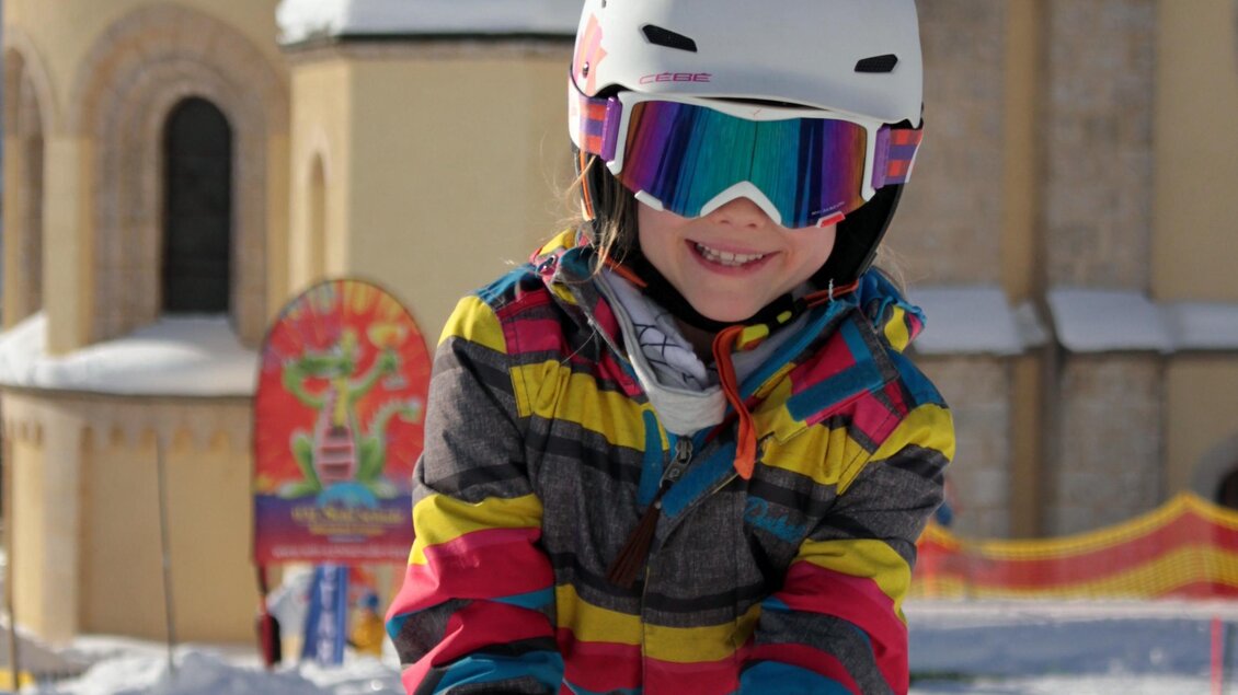 Ein glückliches Kind trägt eine bunte Skijacke und einen Helm. Es steht auf einer Schneepiste vor einer Kirche. | © WM-Schischule Royer