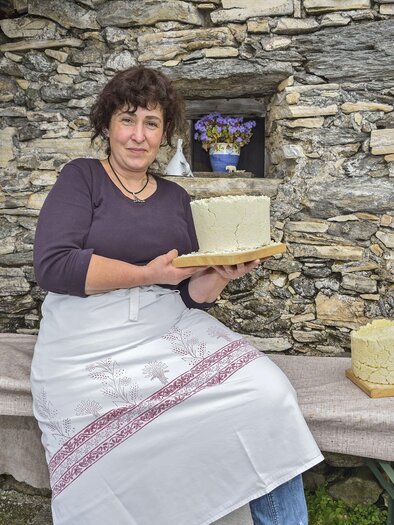 A woman stands in front of a stone house wall holding a cheese in her hands. Next to her is a wooden press and another cheese on the table. | © Herfried Marek