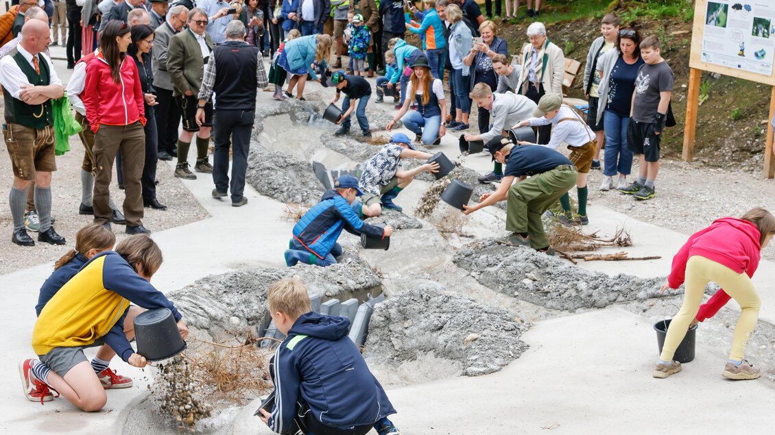 Eine Gruppe von Kindern und Erwachsenen arbeitet zusammen in einer Freiluftumgebung. Sie verwenden Eimer, um Materialien in einem Spielbereich zu bewegen, während Zuschauer zuschauen. | © Martin Huber