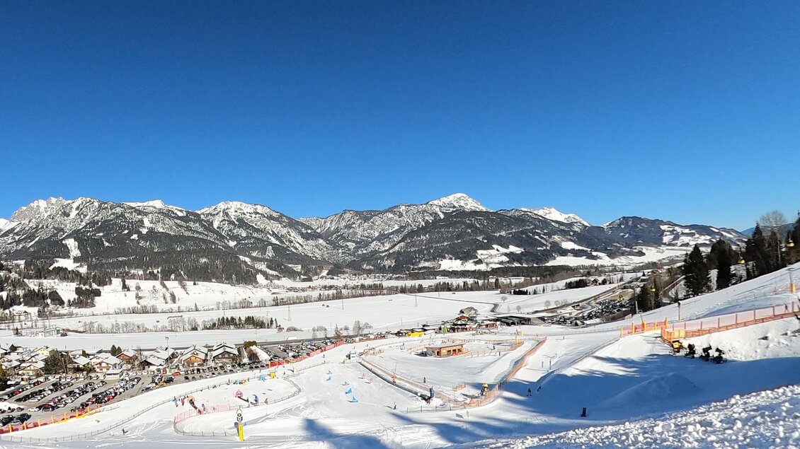 Eine verschneite Landschaft mit Bergen und einem blauen Himmel. Auf der Piste sind Skifahrer und ein Skilift zu sehen. | © Hauser Kaibling