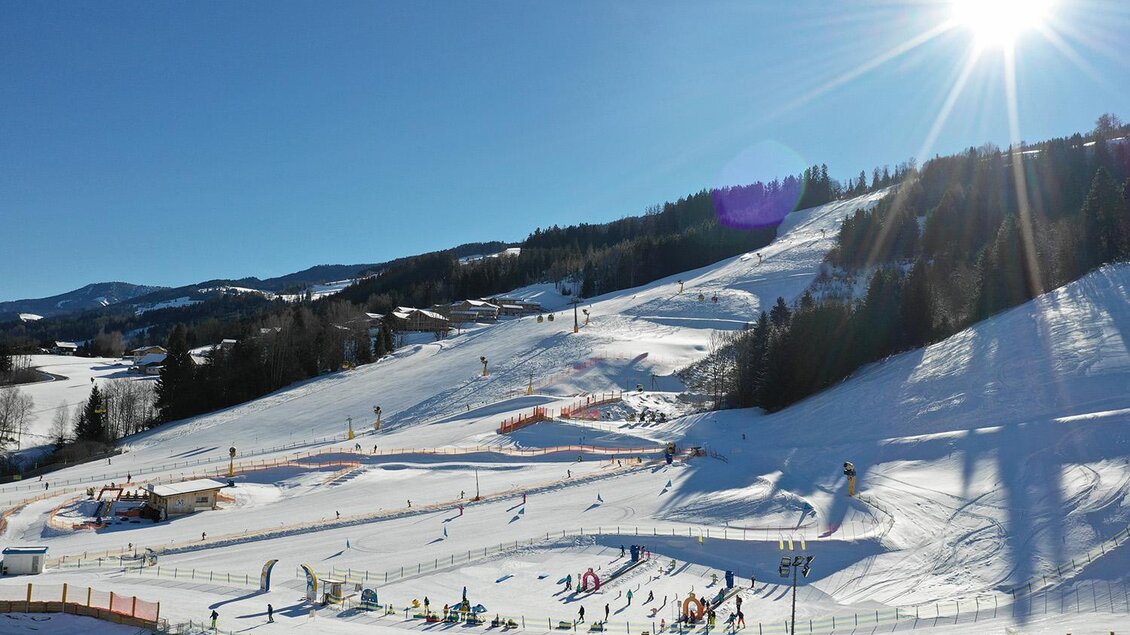 Eine sonnige Skistartrampe mit Schnee und Skifahrern. Im Hintergrund sind bewaldete Hügel und ein blauer Himmel zu sehen. | © Wollis Kids Park