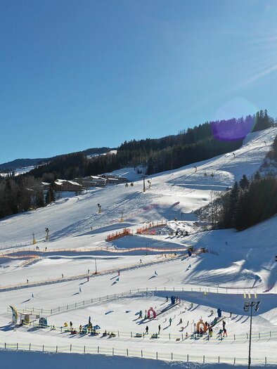 Eine sonnige Skistartrampe mit Schnee und Skifahrern. Im Hintergrund sind bewaldete Hügel und ein blauer Himmel zu sehen. | © Wollis Kids Park