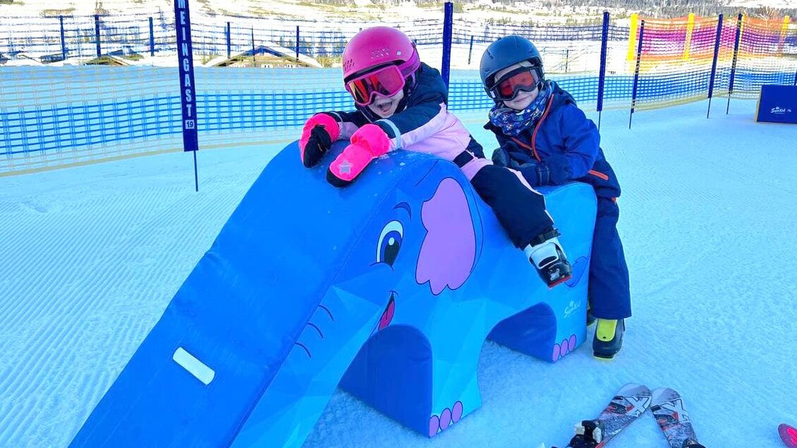 Zwei Kinder sitzen auf einem blauen Elefanten-Spielgerät im Schnee. Im Hintergrund sind Berge und eine schneebedeckte Piste zu sehen. | © Hauser Kaibling