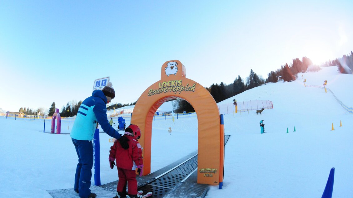 Ein Skilehrer hilft einem kleinen Kind, das bereit ist, die Piste zu betreten. Im Hintergrund sind schneebedeckte Hügel und eine klare blaue Himmel zu sehen. | © Hauser Kaibling