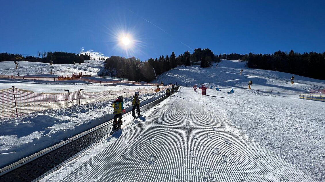 Ein Skigebiet mit schneebedeckten Hängen und einem blauen Himmel. Einige Skifahrer stehen auf einem Förderband. | © Hauser Kaibling