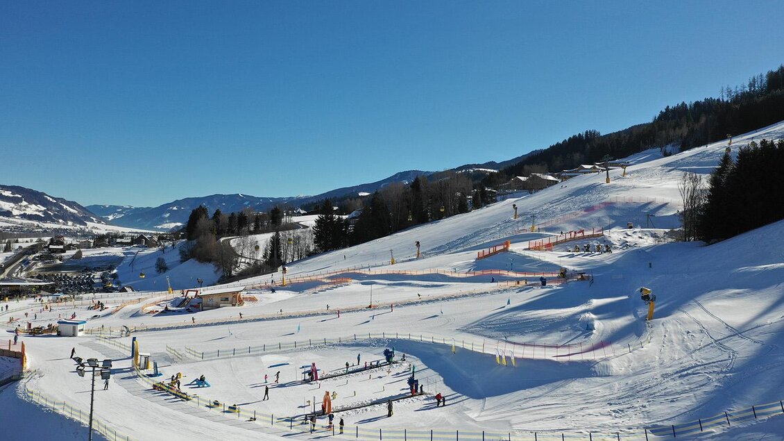 Eine verschneite Wintersportlandschaft mit verschiedenen Skifahrern und Pisten. Der Himmel ist klar und blau, umgeben von Bergen. | © Hauser Kaibling