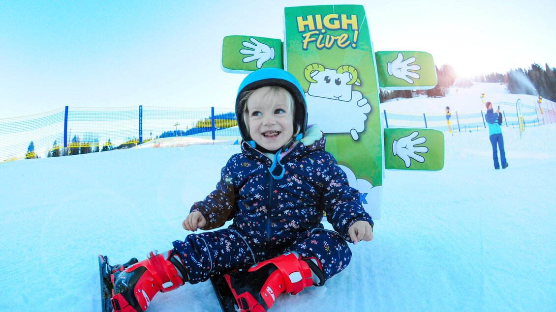 Ein fröhliches Kind sitzt im Schnee und trägt Skiausrüstung. Im Hintergrund steht ein Schild mit der Aufschrift "HIGH FIVE!" und einem Schaf. | © Hauser Kaibling