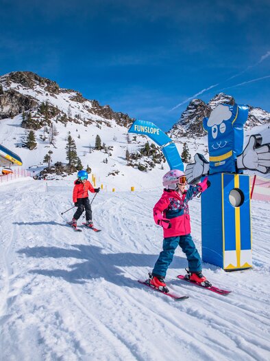 Two children are skiing in a snowy ski resort. In the background is a distinctive figure and the scene is framed by mountains and a blue sky. | © Hauser Kaibling