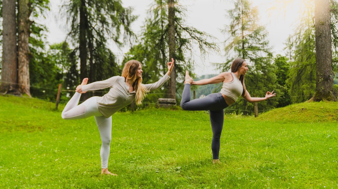 Zwei Frauen üben Yoga im Freien auf einer grünen Wiese. Sie stehen in Positionen, umgeben von Bäumen und einem ruhigen Ambiente. | © Hotel Rösslhof