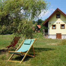 Ein charmantes, gelbes Haus auf einer Wiese. Zwei Liegestühle stehen im Schatten eines Baumes.