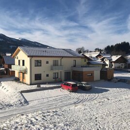 A modern house in a snow-covered landscape. In the background, mountains and other houses can be seen.