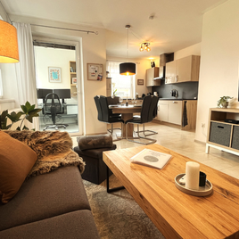 Ein modernes Wohnzimmer mit einem gemütlichen Sofa, einem Holzkaffee Tisch und einem großen Fernseher. Im Hintergrund ist ein Essbereich und eine offene Küche sichtbar. | © Apartment Moritz