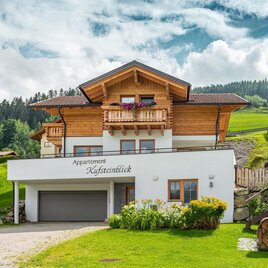 A charming apartment building with a wood and plaster facade, surrounded by green meadows. The tranquil rural setting offers a view of the mountains. | © David Stocker Photography
