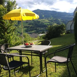 A beautiful garden table with chairs and a yellow sun umbrella. In the background, green hills and a sunny landscape can be seen.