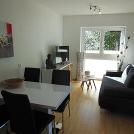 Ein modernes Wohnzimmer mit einem grauen Sofa und einem Esstisch. Große Fenster bieten viel Tageslicht und Blick auf das Grün draußen.