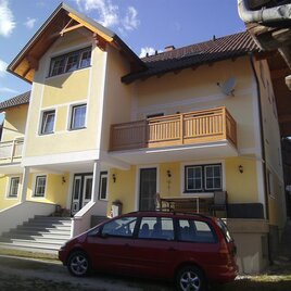 A modern, multi-story residential building with a yellow paint. In front of the house, there is a red car and there is a porch with a wooden railing.