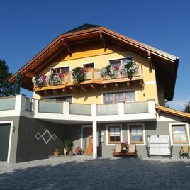 A modern, two-story house with a slanted roof and colorful flowers on the balcony. The entrance is invitingly designed, surrounded by a paved driveway.
