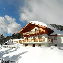Ein schön gelegenes Haus im Schnee mit einem Holzbalkon. Die Umgebung ist von Bäumen und einem klaren, blauen Himmel umgeben.