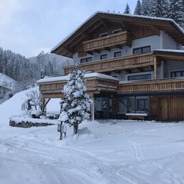 A cozy wooden house in the snow, surrounded by snow-covered trees and mountains. The landscape is wintry and peaceful.