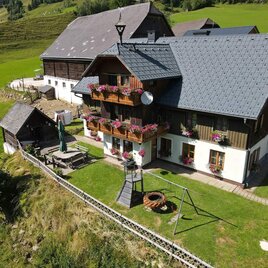 A cozy house in a picturesque landscape, surrounded by green meadows. Colorful flowers bloom on the balcony, and there is an inviting garden area. | © Familie Mörschbacher