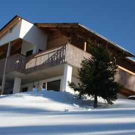 A modern house with wooden cladding stands on a snow-covered hill. The clear blue sky provides a fresh winter atmosphere.