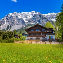 A picturesque house in front of impressive mountains. The surroundings are green and blooming, with a bright blue sky.