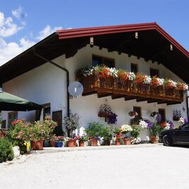 Ein schönes, traditionelles Haus mit blühenden Balkonen und einer gepflegten Gartenlandschaft. Im Vordergrund steht ein schwarzes Auto und ein Sonnenschirm ist aufgestellt.