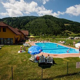 <p>A sunny swimming pool with loungers and sun umbrellas is surrounded by green hills. People are enjoying their leisure time in the water and on the lawn.</p>