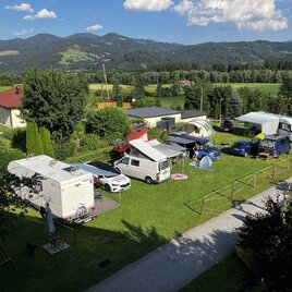 A green meadow with several motorhomes and cars. In the background, mountains and a blue sky can be seen. | © Camping Deutinger