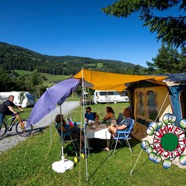 <p>Ein gemütlicher Campingplatz mit einer Gruppe von Menschen, die unter einem Sonnenschirm sitzen, ist zu sehen. Im Hintergrund sind Wohnmobile und eine grüne Berglandschaft.</p>
