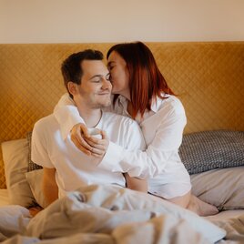 A couple sits on the bed and smiles at each other. The atmosphere is friendly and intimate, with soft pillows and a warm decor. | © Katharina Hafner
