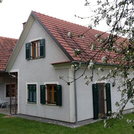 Ein schönes, zweigeschössiges Haus mit rotem Dach und grünen Fensterläden. Der Garten ist von blühenden Bäumen umgeben.