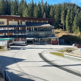 A modern building in the mountains, surrounded by tall fir trees. The road in front of the building is well developed and leads through a picturesque landscape.