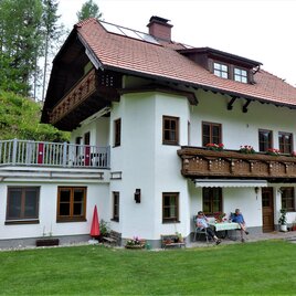 Ein charmantes Haus in den Alpen mit einem gepflegten Garten. Auf der Terrasse sitzen Menschen und genießen die Aussicht.
