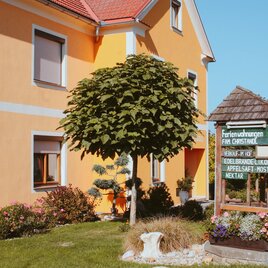 Ein gemütliches Ferienhaus mit orangefarbenen Wänden und einem gepflegten Garten. Im Vordergrund steht ein Schild mit Informationen zu den Ferienwohnungen. | © Christandl