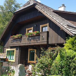 Ein wunderschönes Holzhaus mit einem begrünten Garten. Im Hintergrund sind Berge und ein blauer Himmel sichtbar.