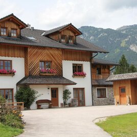 A typical Alpine house with wooden elements and blooming window boxes. In the background, green mountains and a clear sky can be seen.