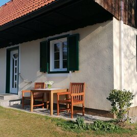 Ein gemütliches Haus mit grünen Fensterläden und einem kleinen Tisch mit Stühlen im Vordergrund. Der Garten ist gut gepflegt und die Sonne scheint. | © Zmugg
