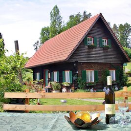 Ein malerisches Haus mit einem roten Dach, umgeben von einer grünen Landschaft. Auf einem Tisch im Vordergrund stehen eine Flasche Wein und Gläser. | © Ferienhaus Possath | Familie Possath